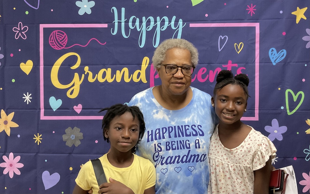 Three individuals stand close together, smiling, with a banner in the background that reads "Happy Grandparents."