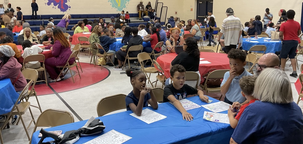 A large group of people in a gym, seated at tables with blue tablecloths, some with papers.