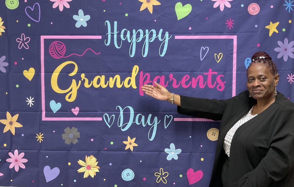 A woman in a black suit stands before a banner with "Happy Grandparents Day" in colorful letters and hearts.