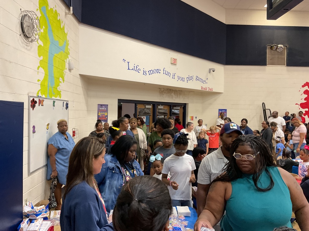 A room with tables full of items; a group of people stand around talking. A sign reads "Life is more fun if you give".