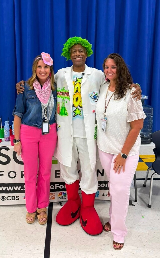 A man in a white jacket and green wig stands with two educators