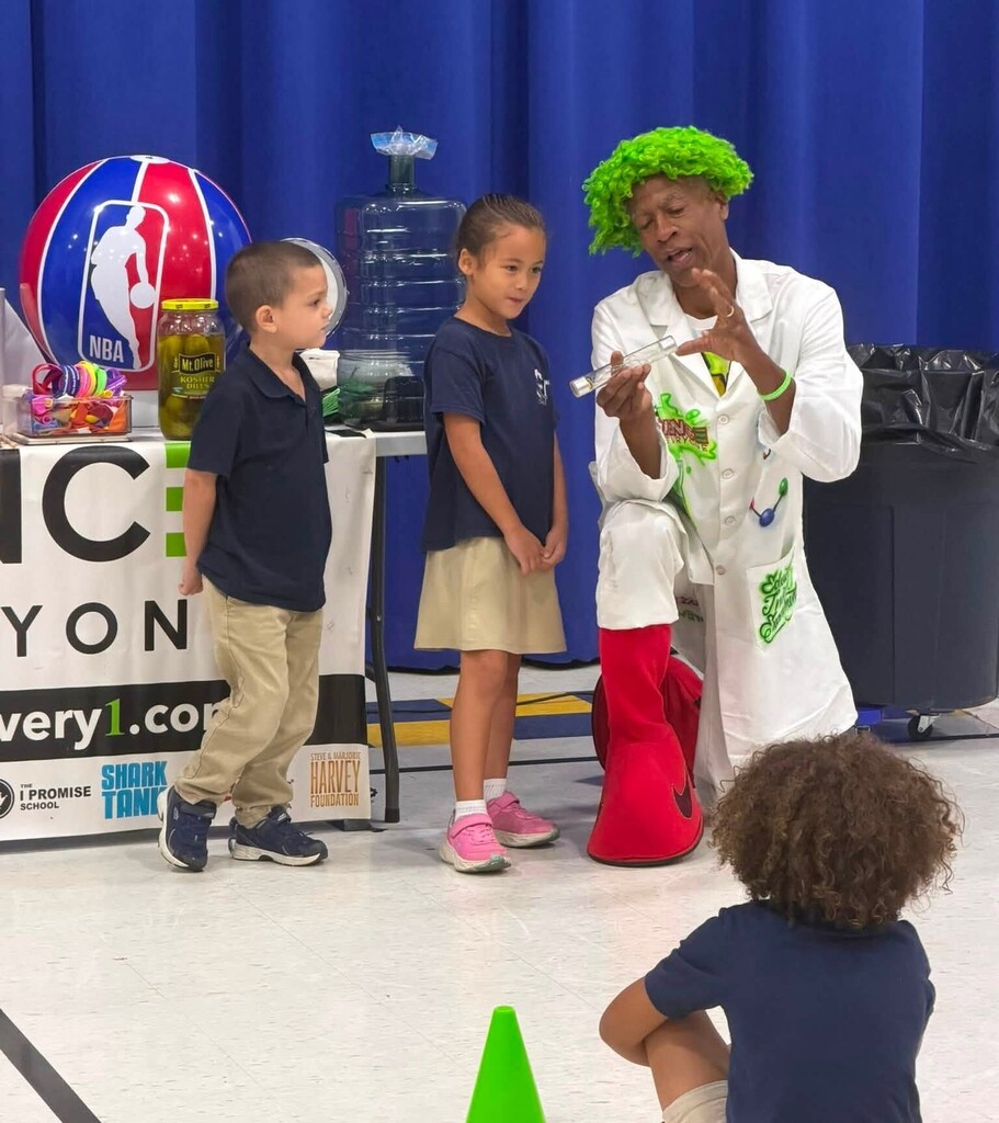 A man in a white jacket and green wig shows a test tube to two students