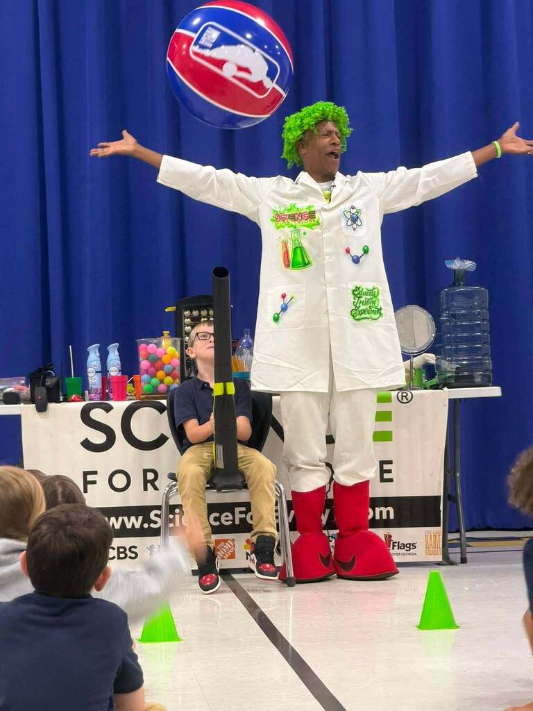 A man in a white jacket and green wig stands with arms outstretched as a student sits in a chair with a blower and a beach ball hovers in the air