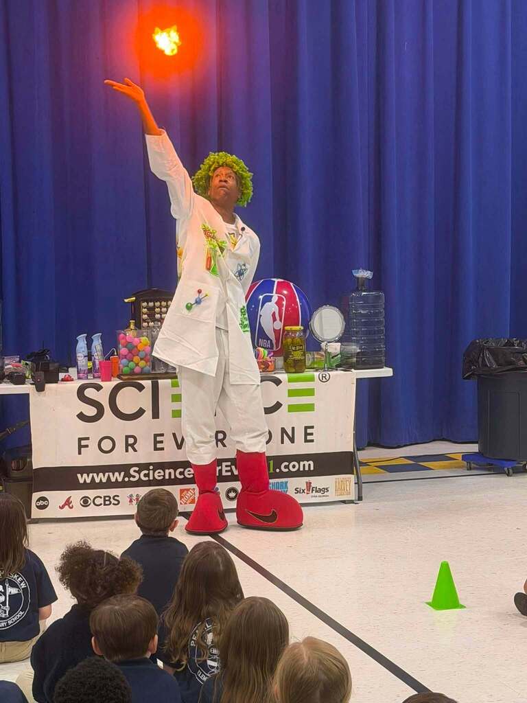 A man in a white jacket, red oversized shoes and a green wig points to a small fireball above him