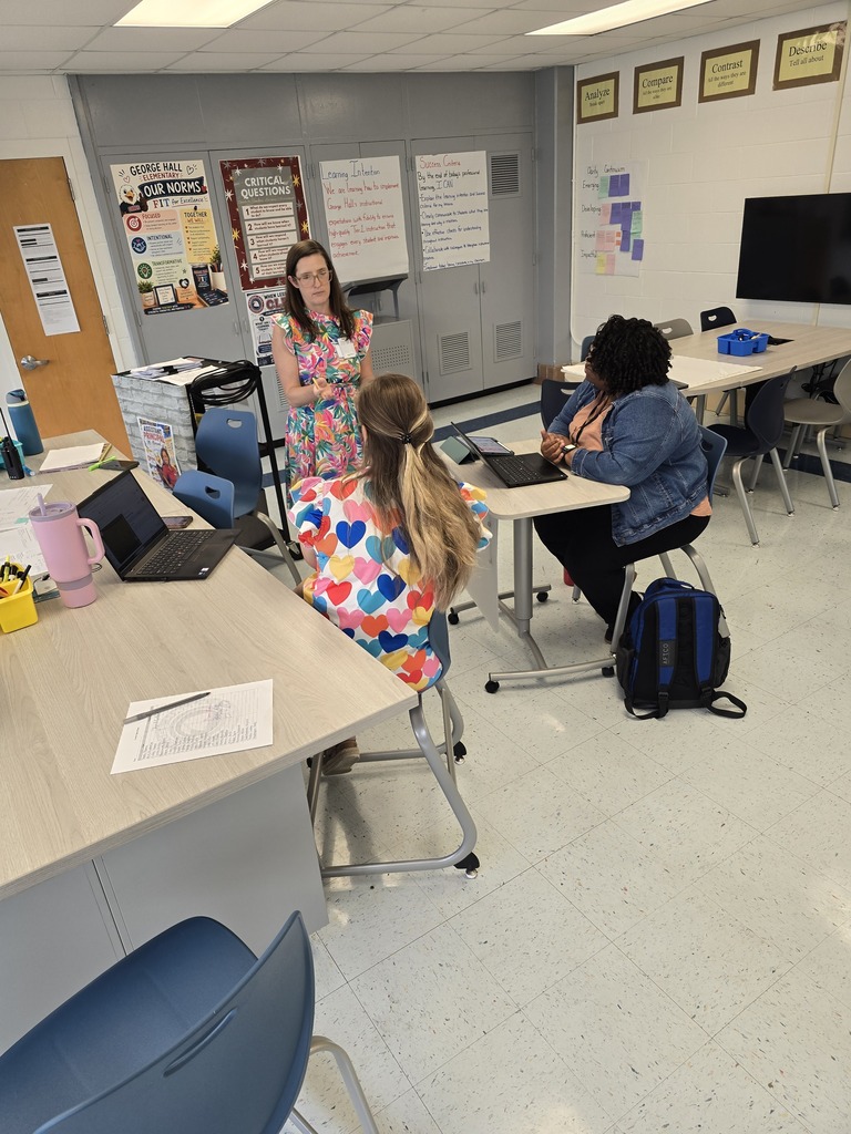 A classroom with desks and chairs, a teacher standing, and two students sitting at desks with laptops.