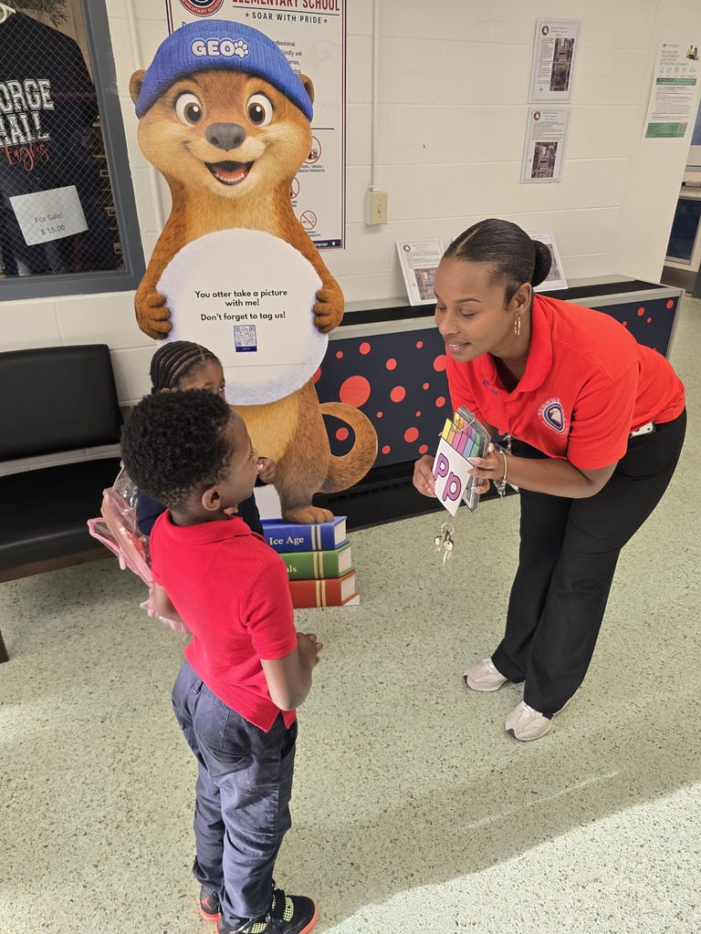 An adult in red hands a boy in red a book. A child stands behind them, near a large, furry mascot.