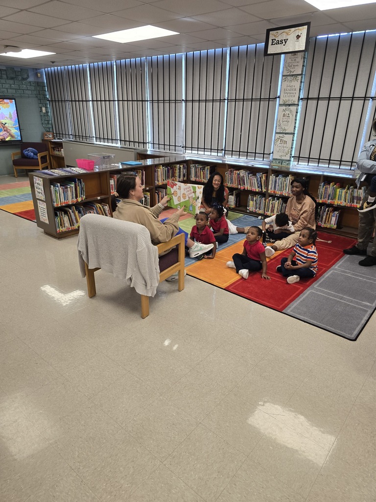 A library with bookshelves. A person reads a book to children on a rug while others sit nearby.