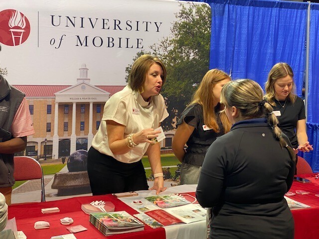 A woman in front of a University of Mobile banner talking with a student