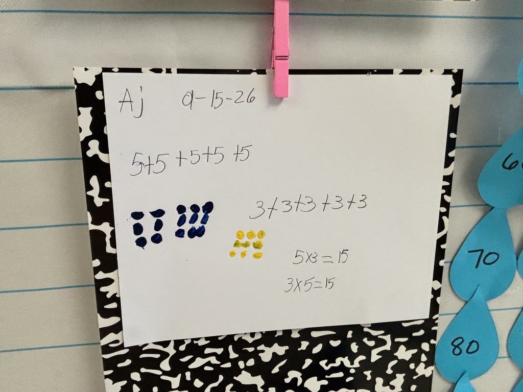A white paper displays various math problems, with a black and white patterned border. Next to it, blue droplets with numbers 60, 70, and 80 hang on the wall.