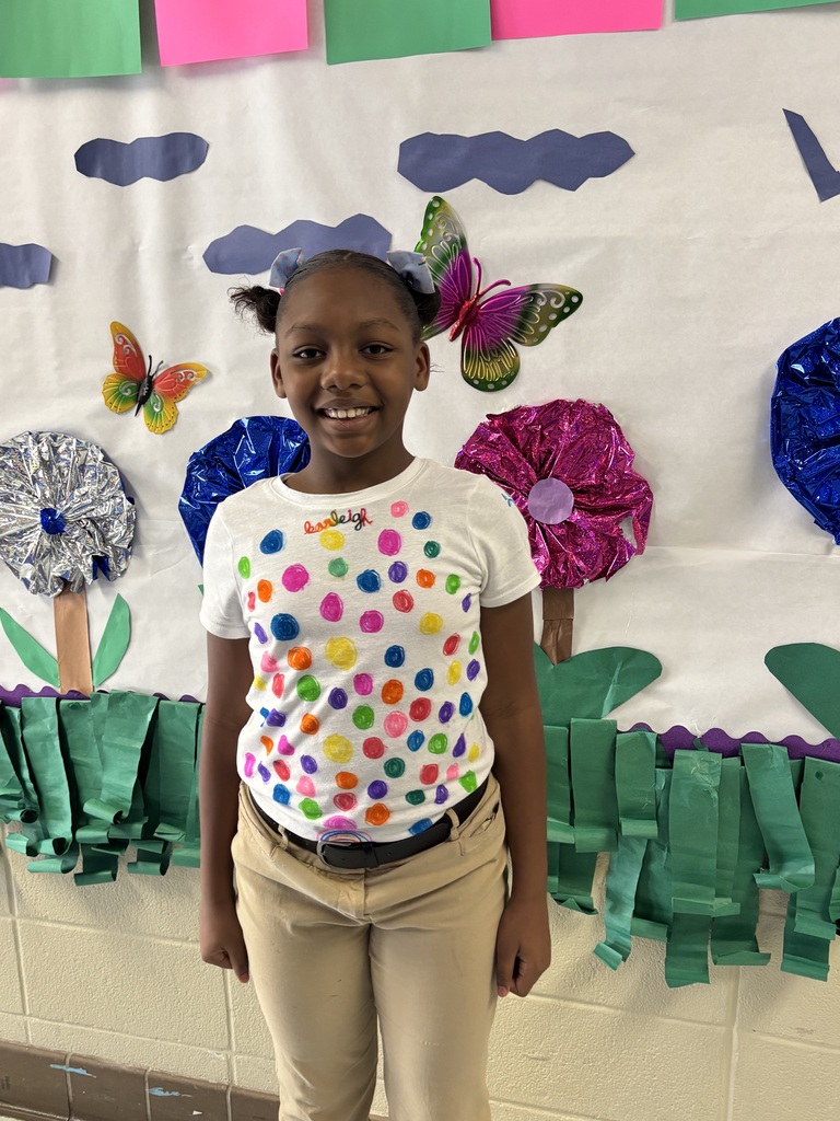 A young girl stands smiling in front of a classroom backdrop with colorful butterflies, flowers, and a green border.