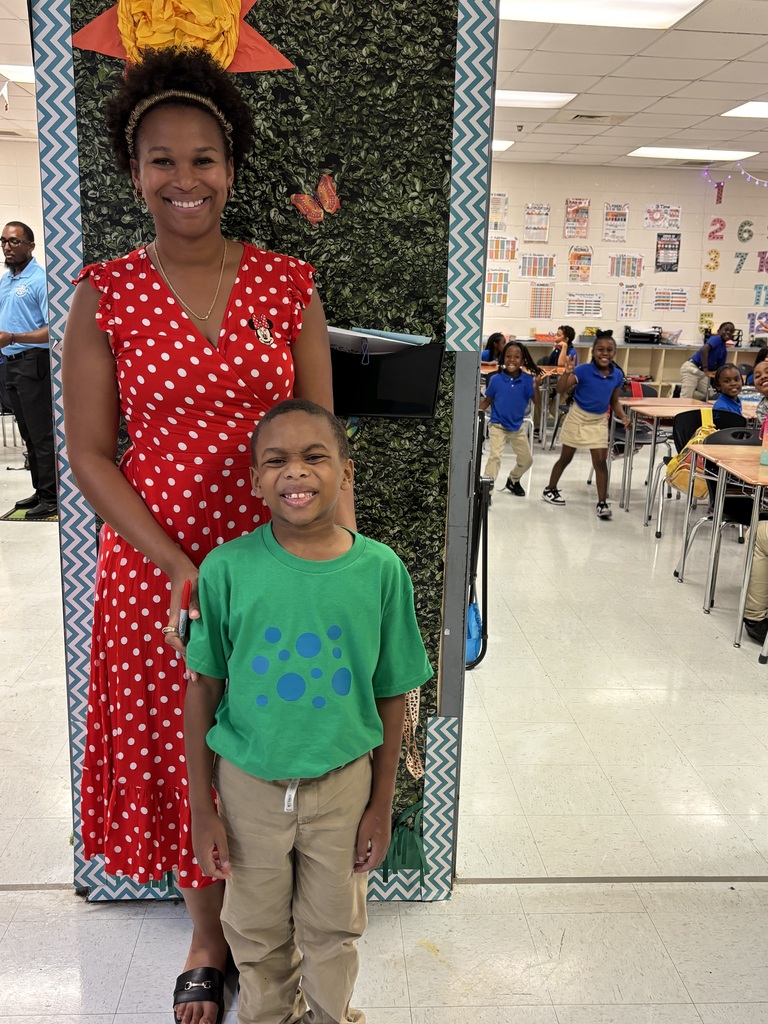 A woman in a red dress stands with a boy in a green shirt in a classroom.