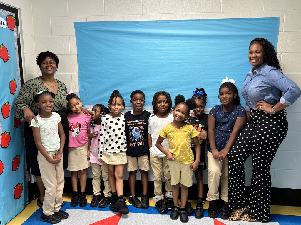 A group of children and two adult women stand in a classroom. They wear various outfits. A blue backdrop with red apples is behind them.