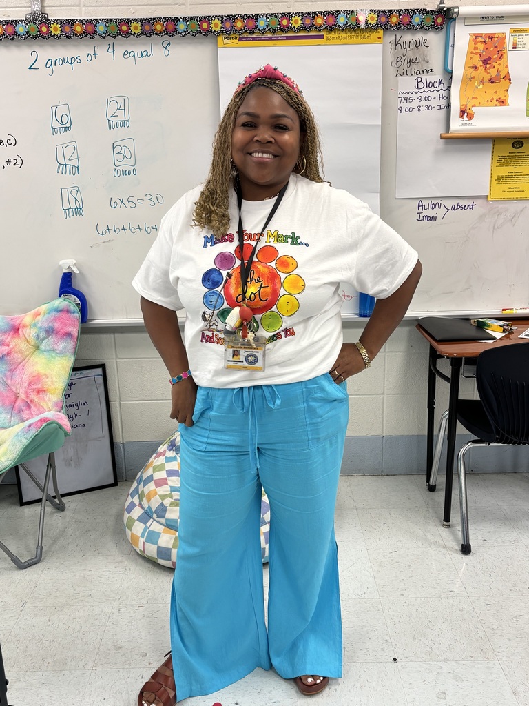 A woman in a white T-shirt and blue pants stands in a classroom. Behind her, a whiteboard displays a list and diagrams.