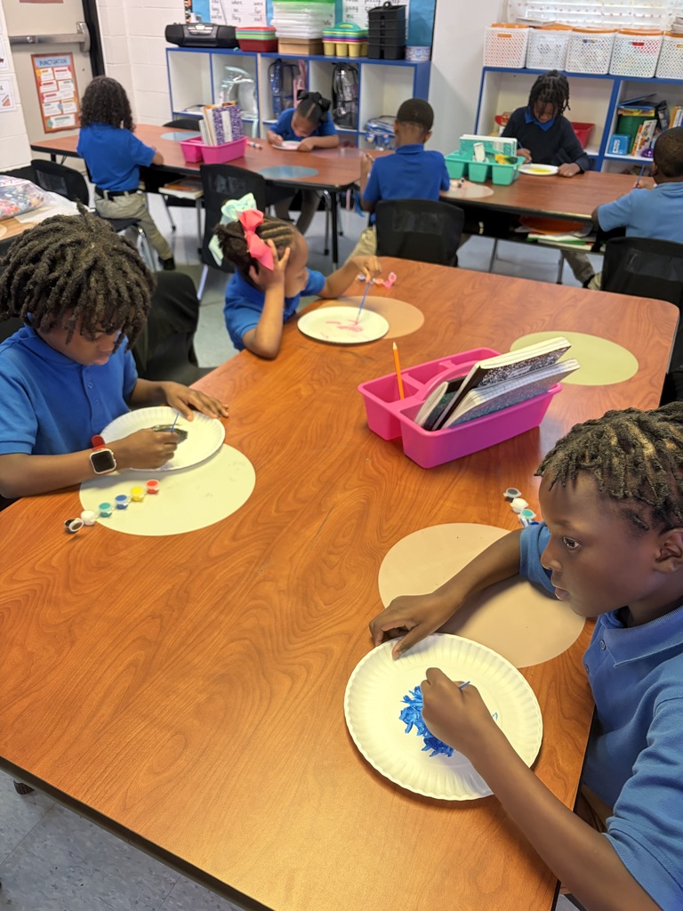 Students at a table, engaged in an activity with plates and art materials. Desks and chairs are in a classroom setting.