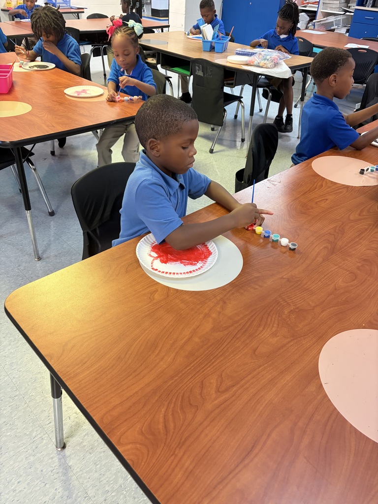 A classroom with children sitting at desks, wearing blue shirts. A child in the front is painting a paper plate.