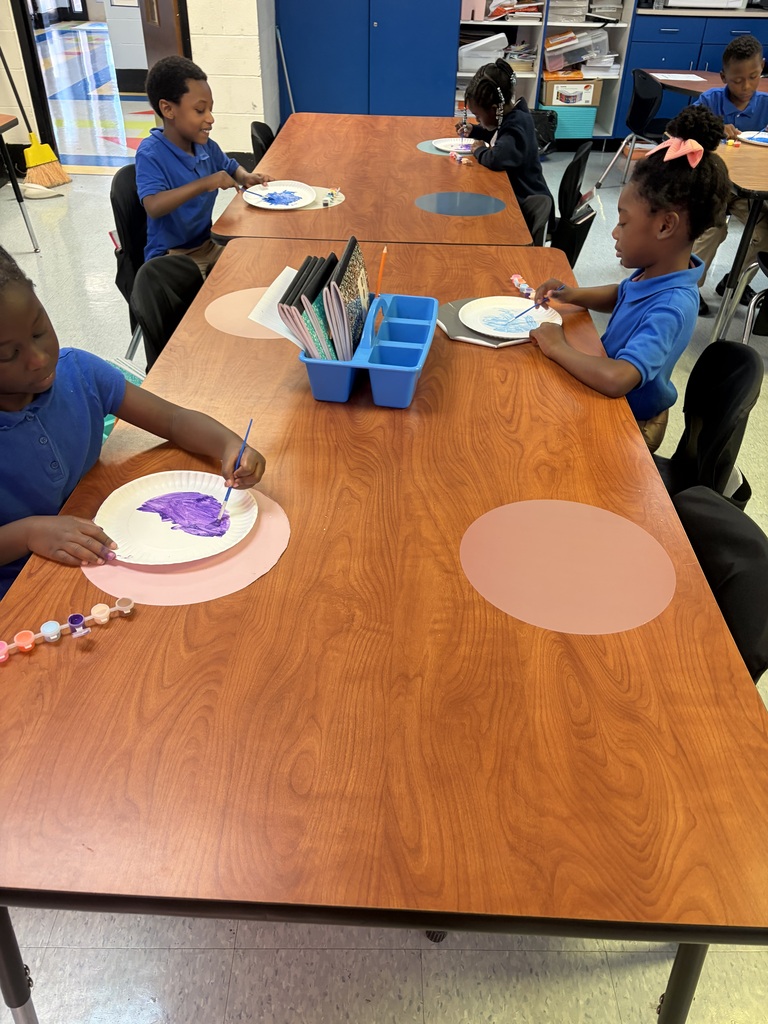 Several children at desks, painting on circular paper with paint. A cabinet and other desks in the background.