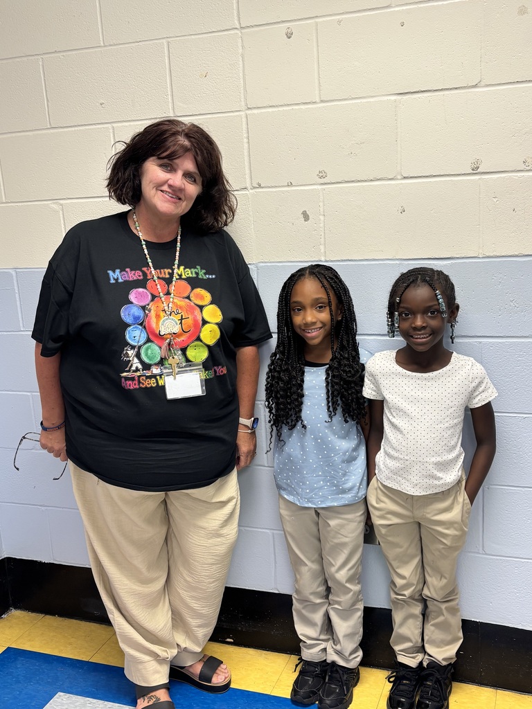 A woman and two young girls stand together in front of a white brick wall. The woman wears a black shirt, while the girls wear blue and white shirts.