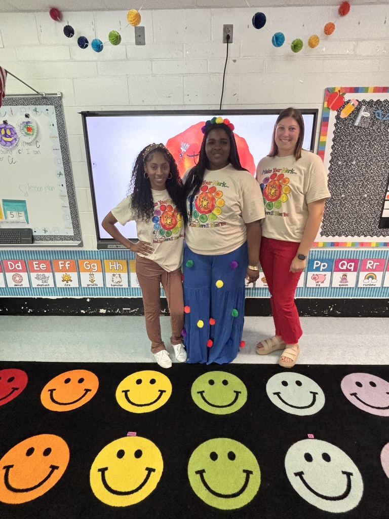 Three women in matching t-shirts stand on a carpet with smiley faces in a classroom.