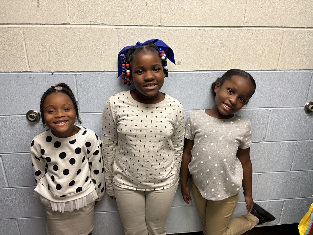 Three girls stand close together against a white wall, smiling. Each wears a different colored top with polka dots.