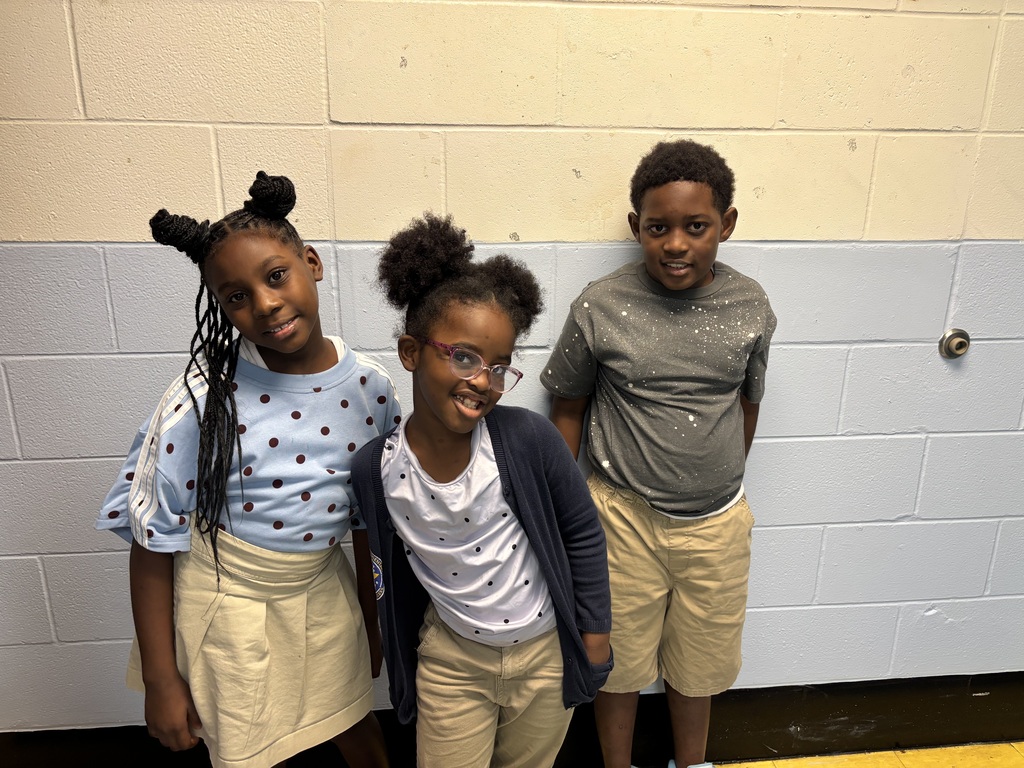 Three children stand together in front of a white wall. They wear casual clothing. One has glasses.