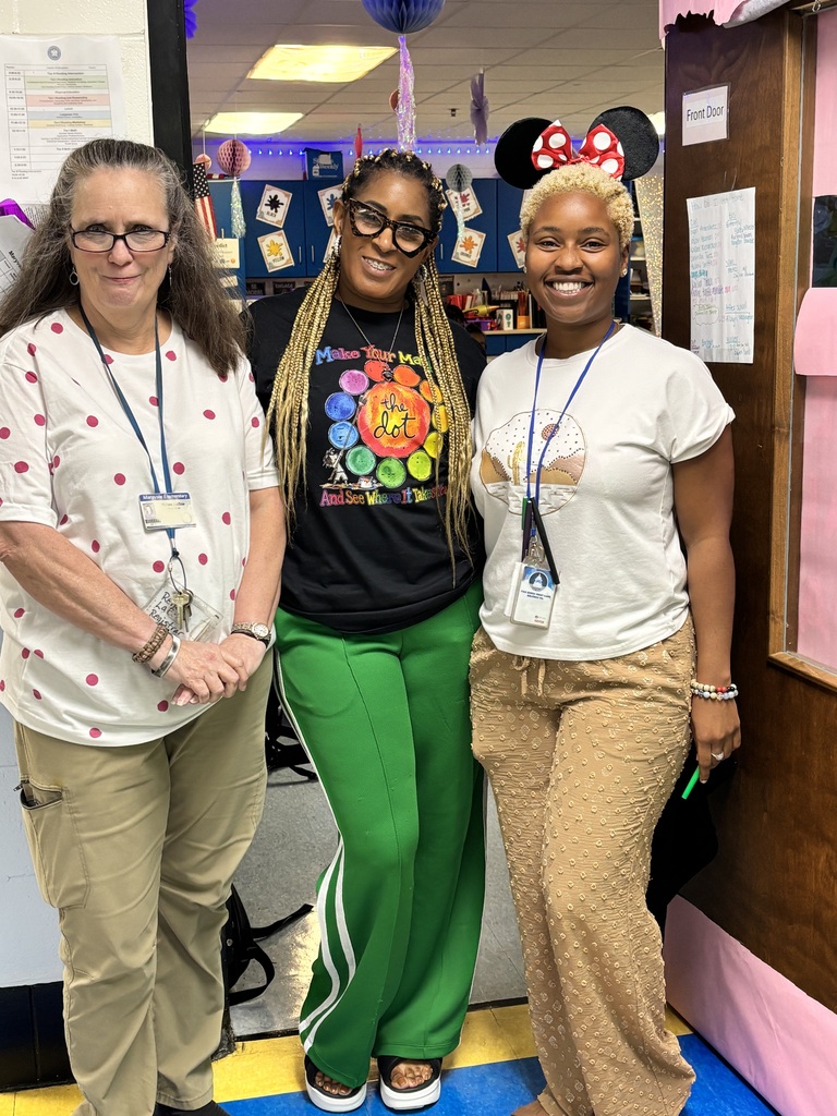 Three women in casual clothing, standing next to each other. One wears glasses, another wears a Mickey Mouse headband. Background shows a door and bulletin board.