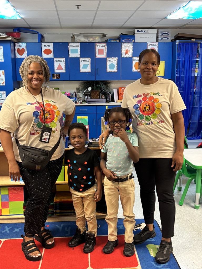 Three adults and two children stand together in a classroom, on a colorful rug. The adults wear beige shirts. The children wear glasses, a black shirt, and khaki pants.