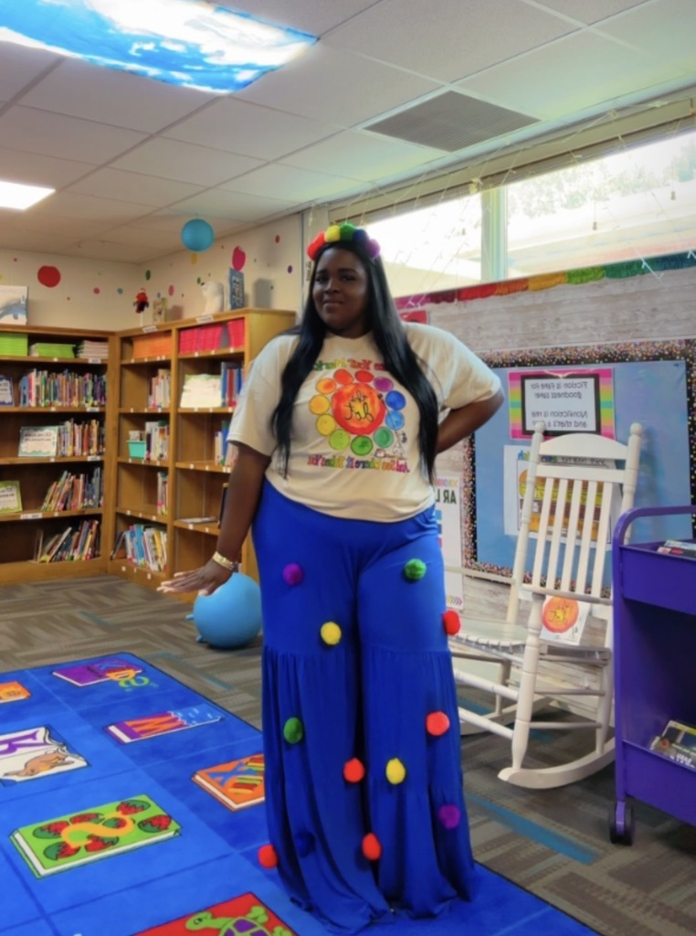 A person in a library, wearing a white shirt, blue pants with colorful pom-poms, and standing on a blue carpet.