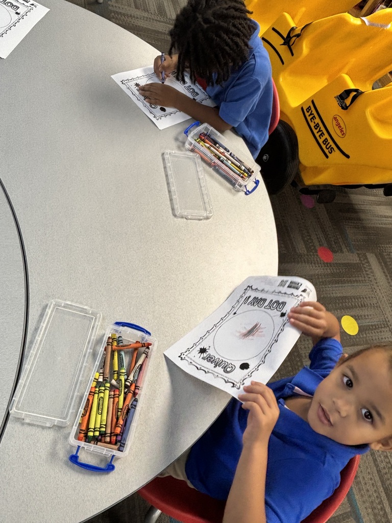 A child holds a paper with a clock face while sitting at a table. Another child sits nearby, working on a paper. Both wear blue shirts. The table has crayons and clear containers.