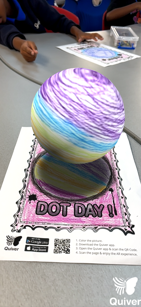 A colorful, multi-layered sphere with swirling patterns is displayed on a table with a printed sheet. It says "Dot Day!" with a reflection below.