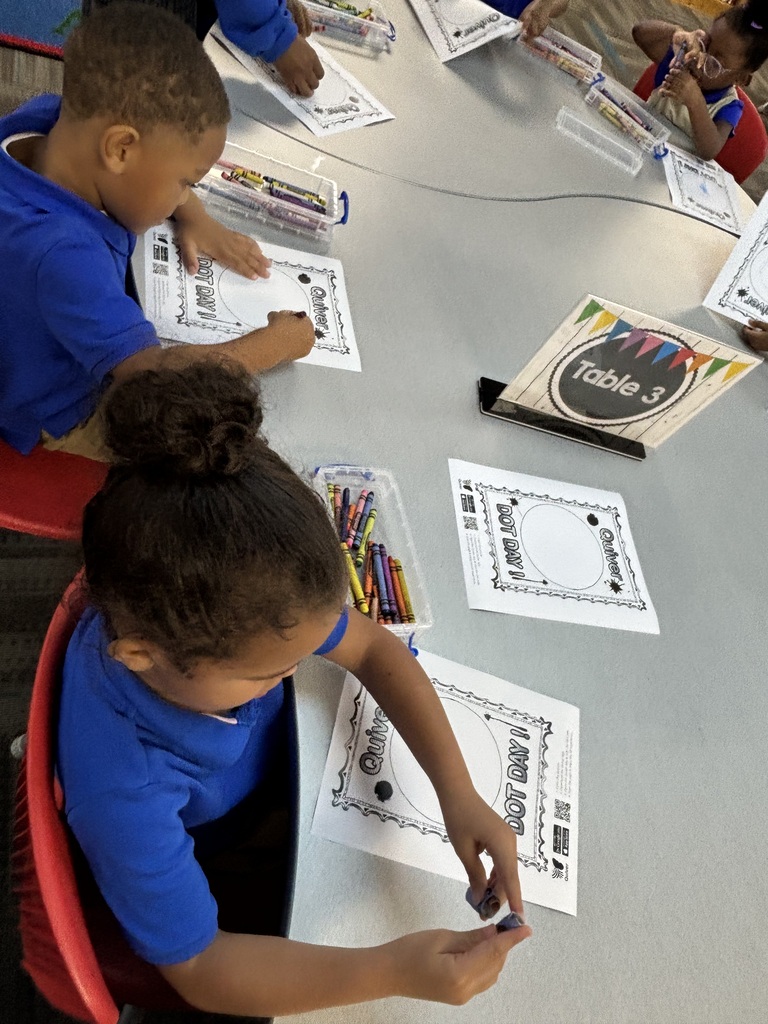 Children in blue shirts sit at a table, engaged in coloring and reading activities. Papers and crayons are on the table.