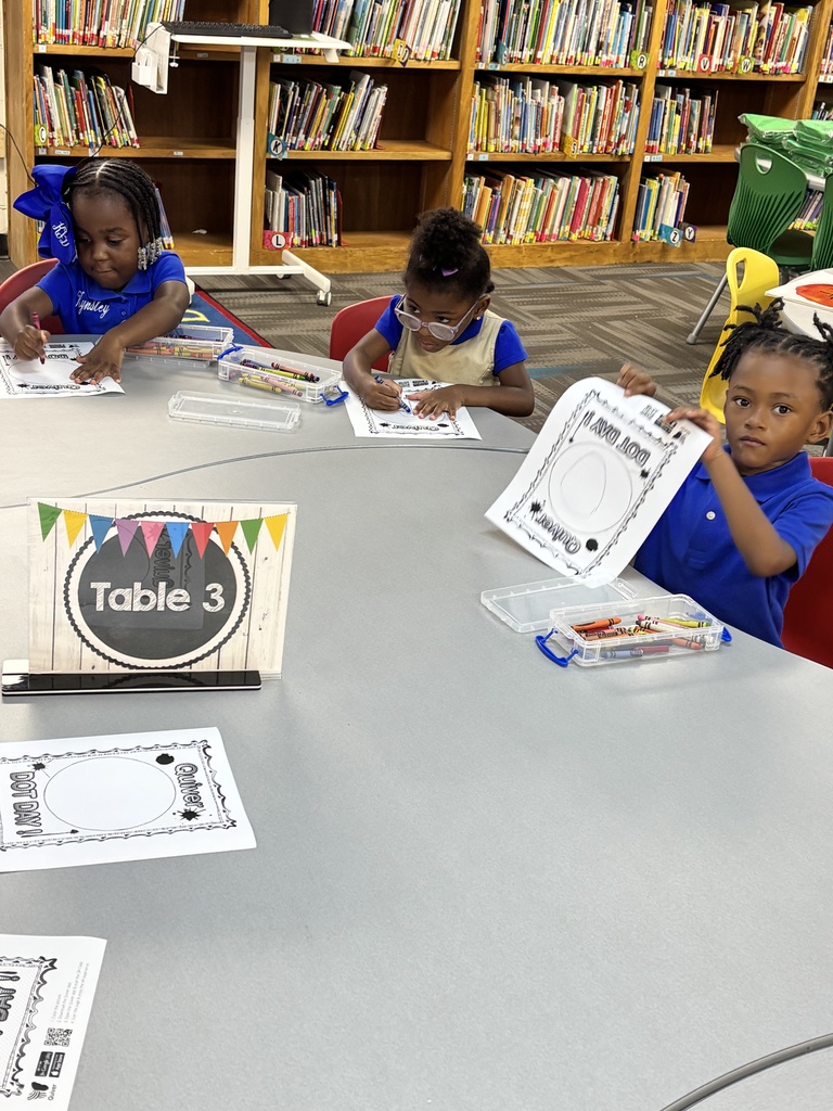 Three children sit at a library table, each with papers and pencils. Shelves with books are in the background.