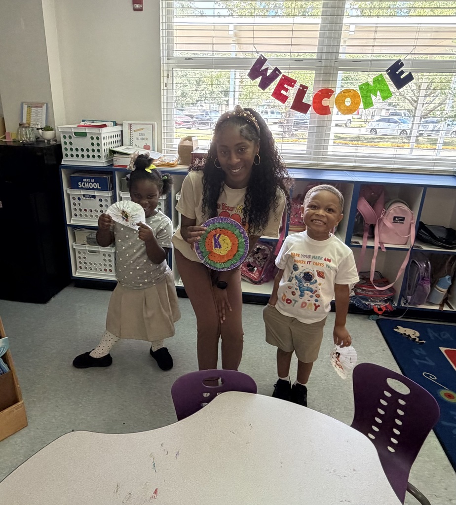 A woman stands with two children in a classroom. She holds a circular object, while the children stand on either side of her.