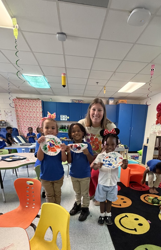 Three children holding paper crafts and a woman stand in a classroom. Behind them are tables, chairs, and a floor mat with smiley faces.