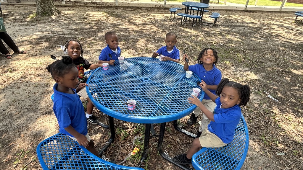 Six children wearing blue shirts sit around a blue picnic table in a park, with a tree and benches in the background.