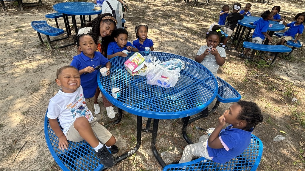 A group of children in blue uniforms sit around blue picnic tables in an outdoor area.