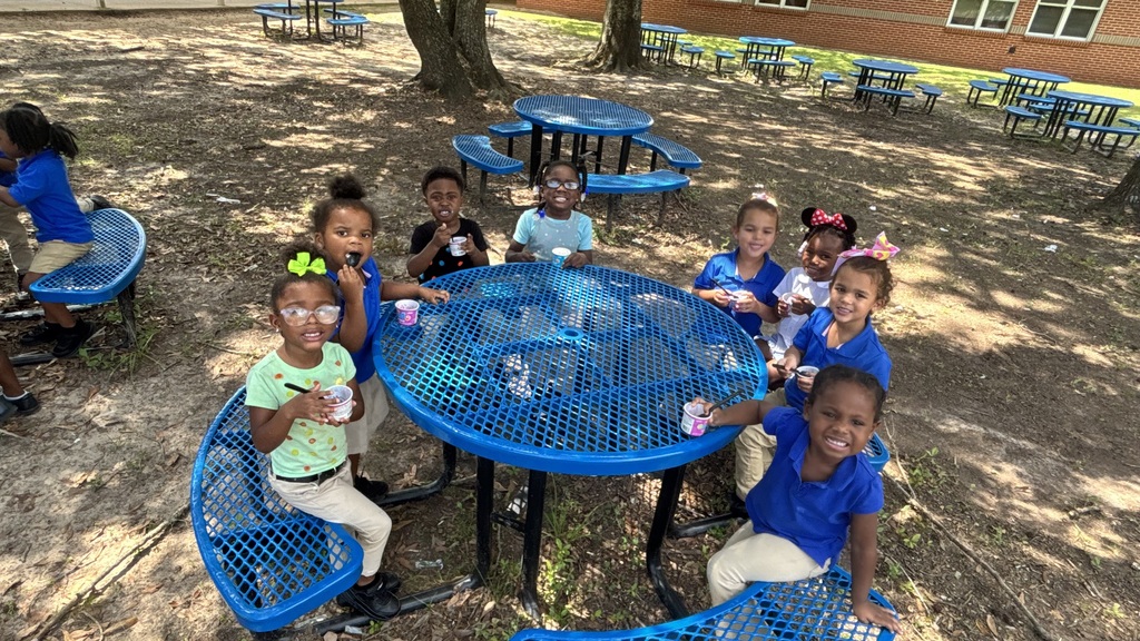 Several young children sit around a blue picnic table, smiling and holding ice cream cups. They wear blue shirts.