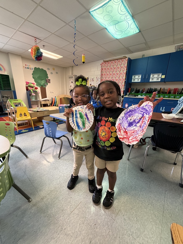 Two children in a classroom with art projects, desks, chairs, and a ceiling light. One holds a white paper creation; the other holds a colorful, abstract one.