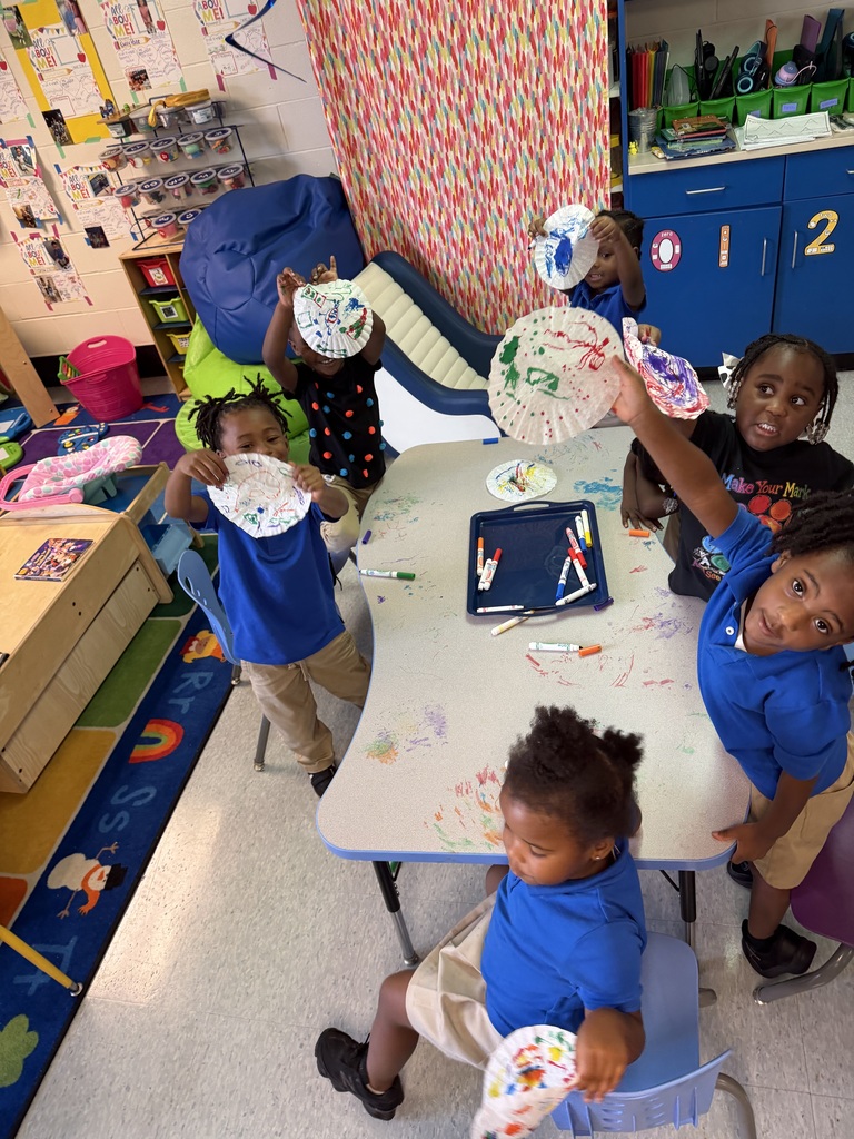 Several children in blue shirts seated at a table with paper plates, holding and displaying their artwork.