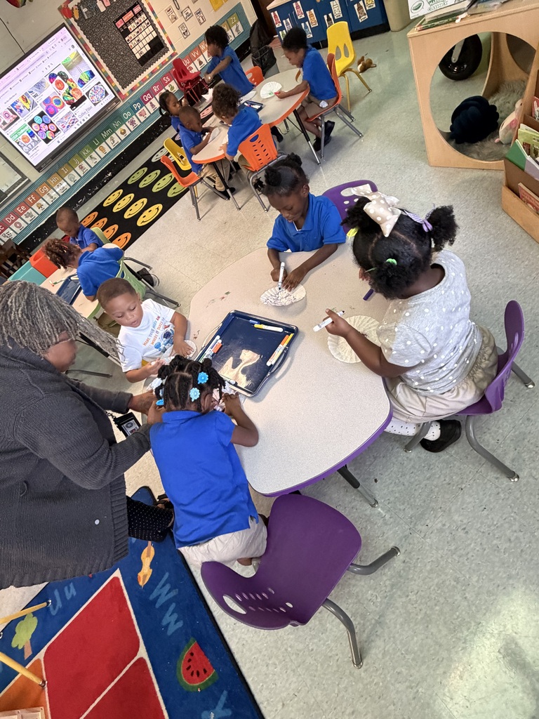 Children in blue uniforms sit at tables in a classroom with an adult, engaging in an activity.