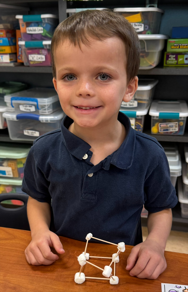 A child sits at a table with a small triangular structure made of marshmallows and toothpicks.