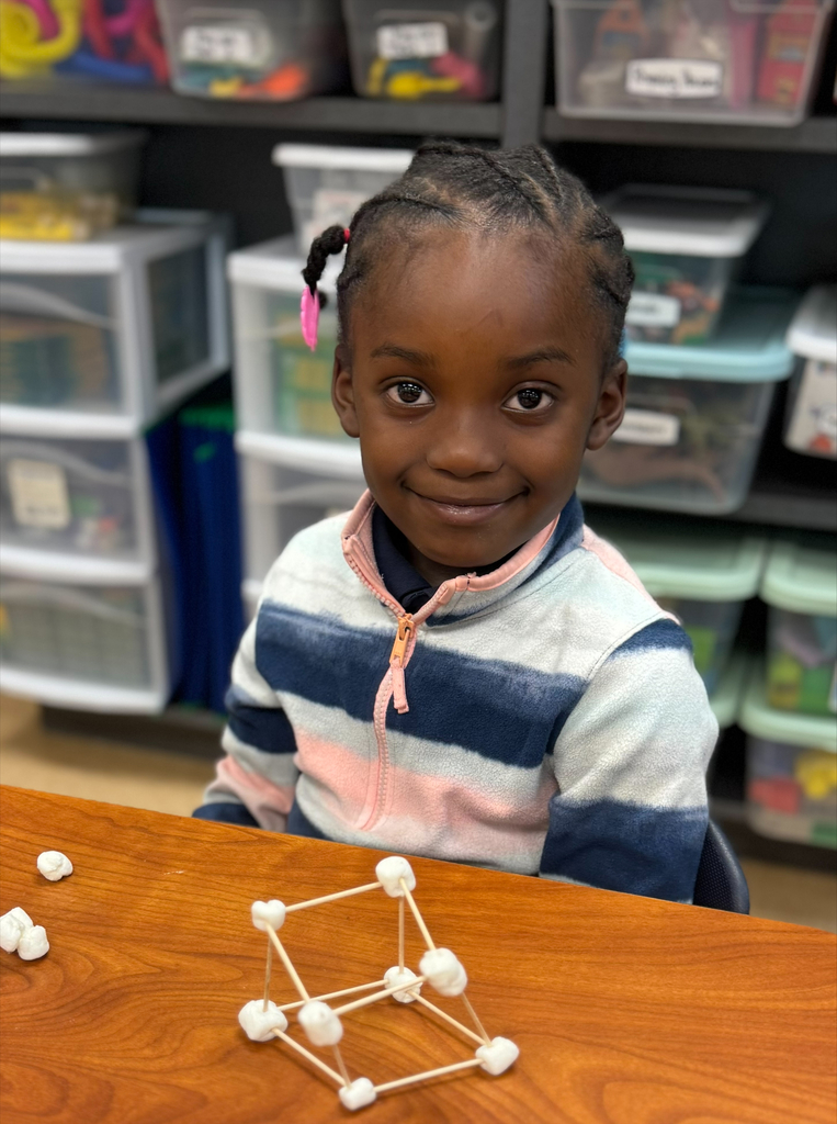 A young girl sits at a table with a model made of marshmallows and toothpicks.