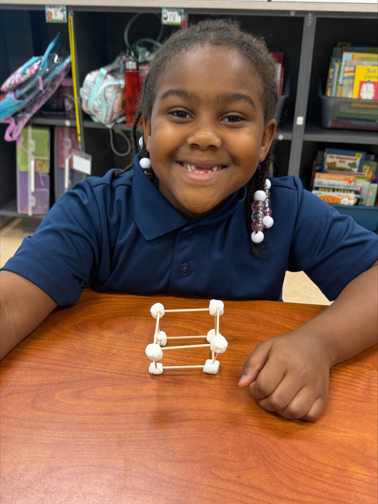 A girl in a blue shirt smiles at the camera while sitting at a desk with a small marshmallow structure.
