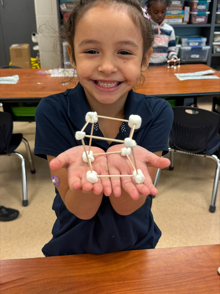 A young girl in a blue shirt holding a triangular structure made of marshmallows and toothpicks.