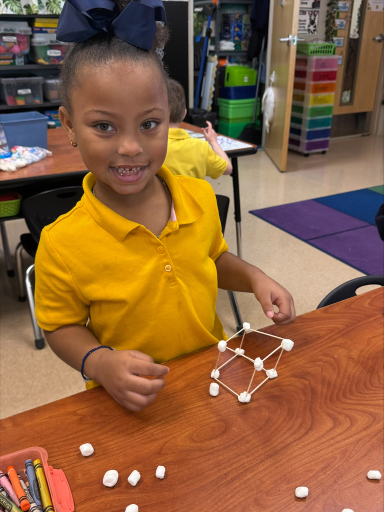 A young girl wearing a yellow shirt builds a structure with marshmallows and toothpicks at a classroom table.
