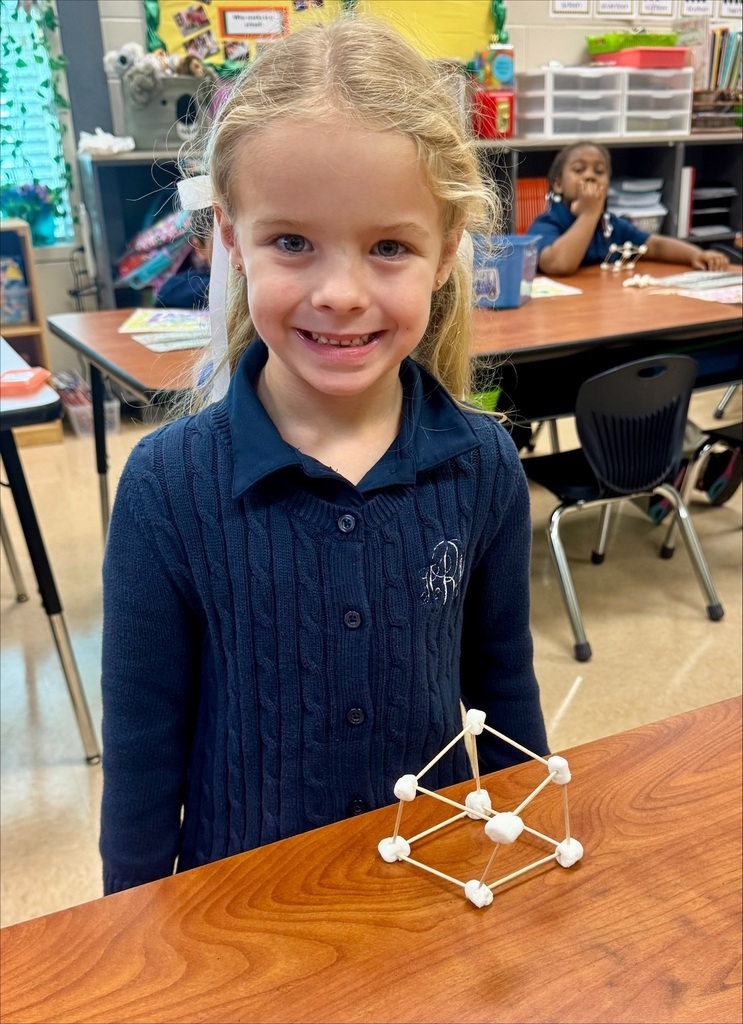 A young girl in a blue shirt stands at a table, smiling while showing a model she built.