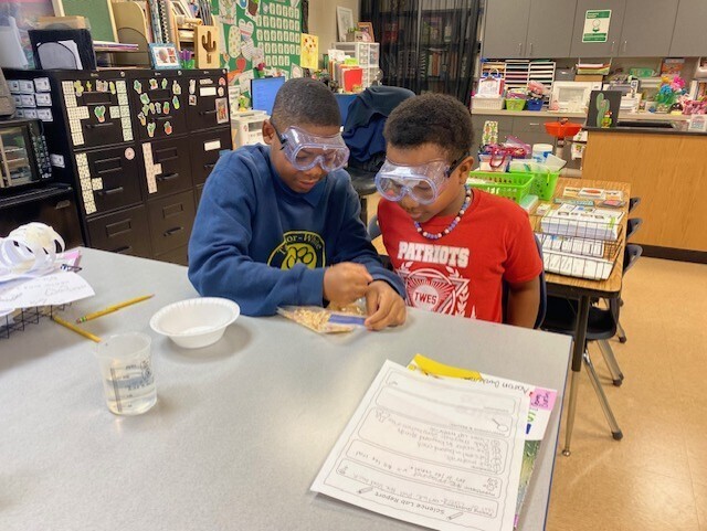 Two students with safety goggles examine a paper and a bowl of liquid on a table.