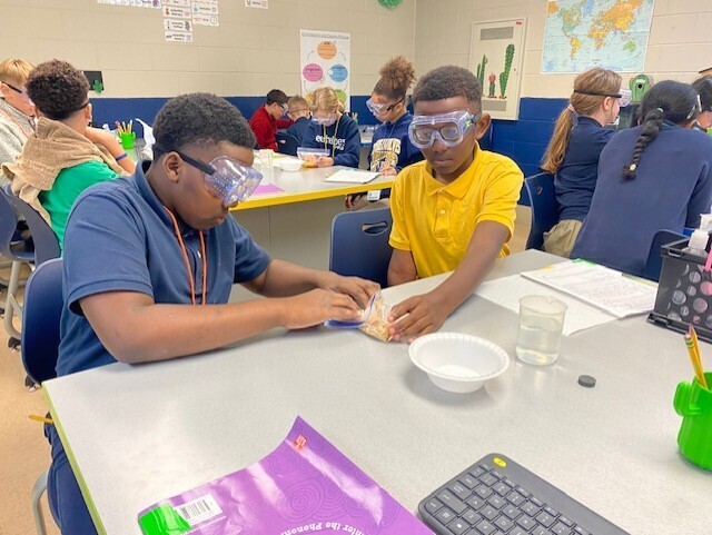 Students wearing goggles work on an activity at desks in a classroom. A table has a bowl, a glass, a keyboard, and papers.
