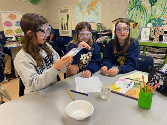 Three students with safety goggles conduct an experiment at a table, holding papers and a container.