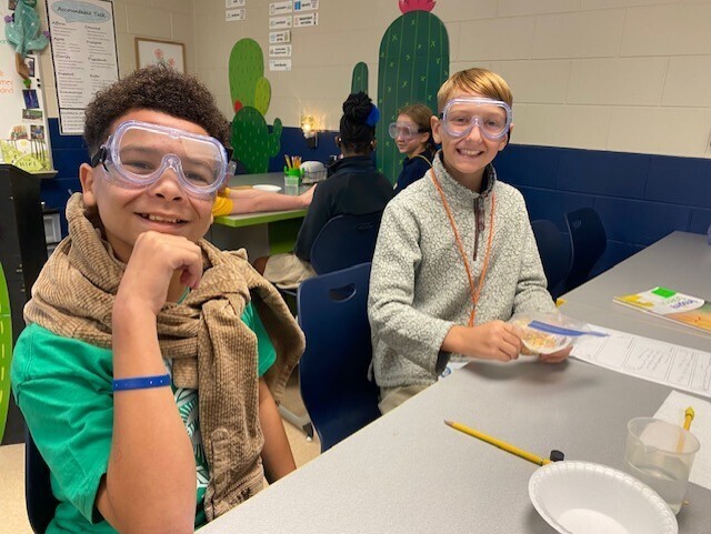 Two kids wearing goggles and scarves sit at a desk with a bowl, papers, and a pencil.