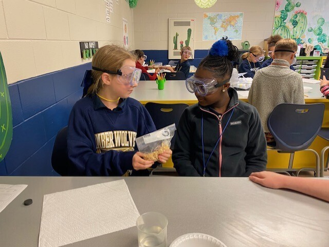Two students wearing goggles work at a table. One holds a plastic bag with food. Behind them, more students sit at tables.